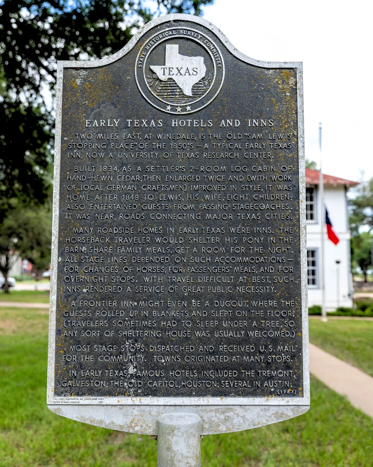 A Texas state historical marker titled Early Texas Hotels and Inns, standing on a green lawn with a white building and a Texas flag behind it.