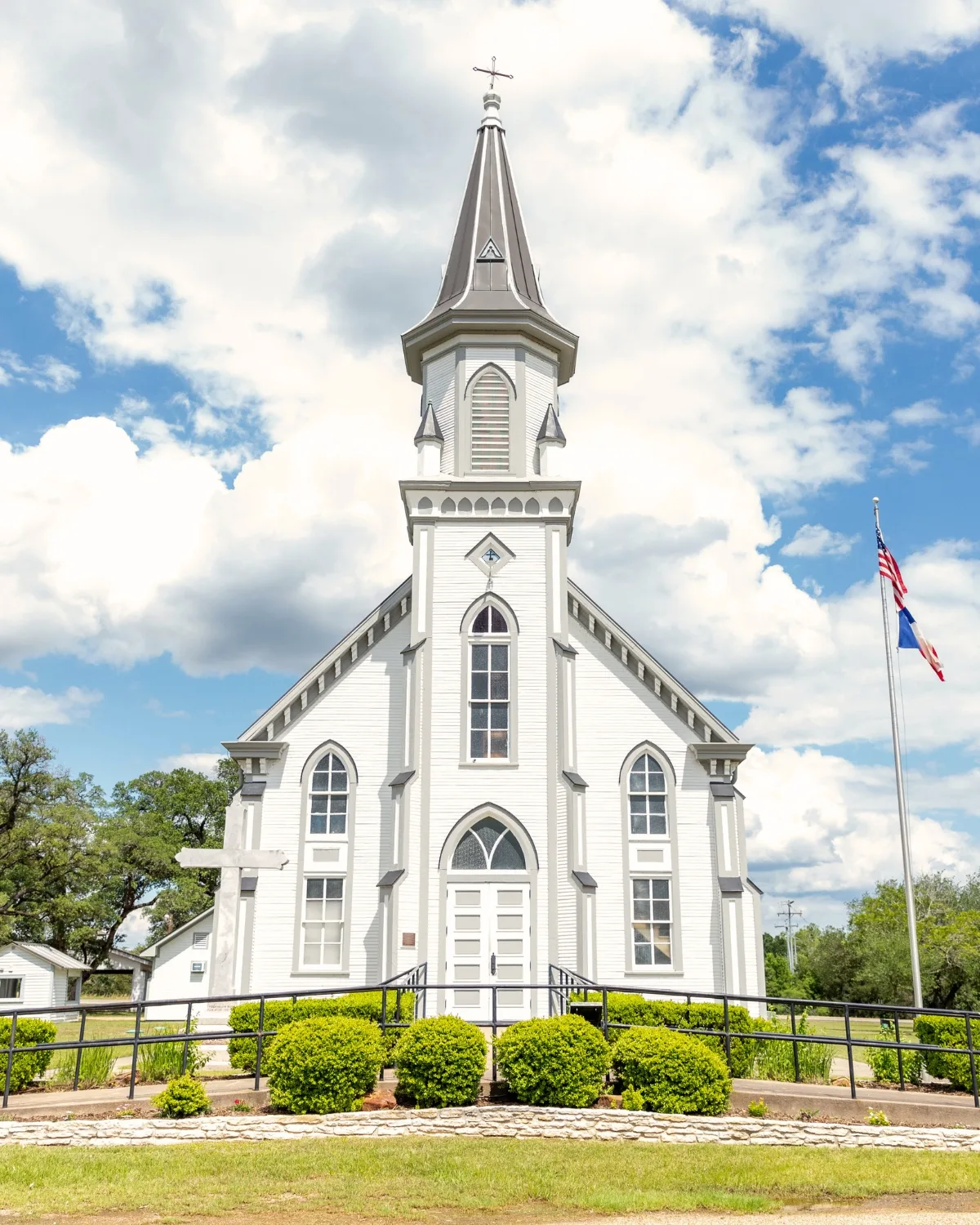 A white clapboard country church with a tall gray steeple, arched stained-glass windows, and clipped hedges along the front walk under a blue sky with tall clouds.