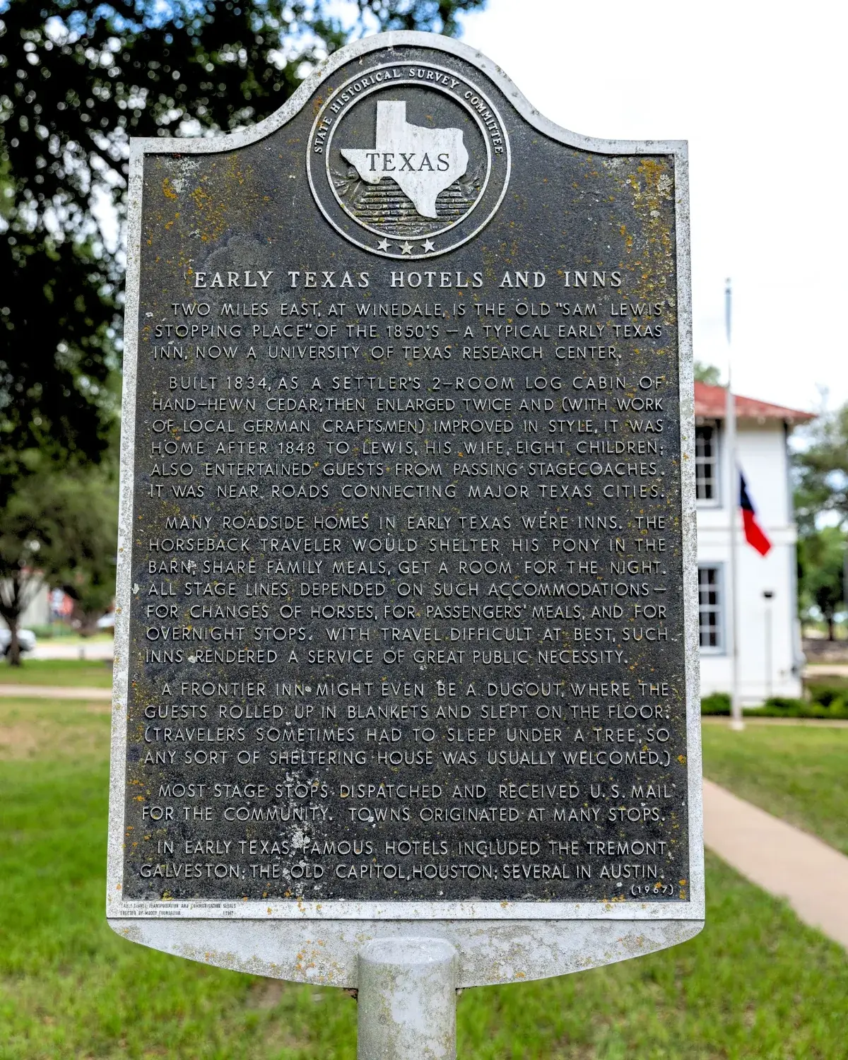 A Texas state historical marker titled Early Texas Hotels and Inns, standing on a green lawn with a white building and a Texas flag behind it.