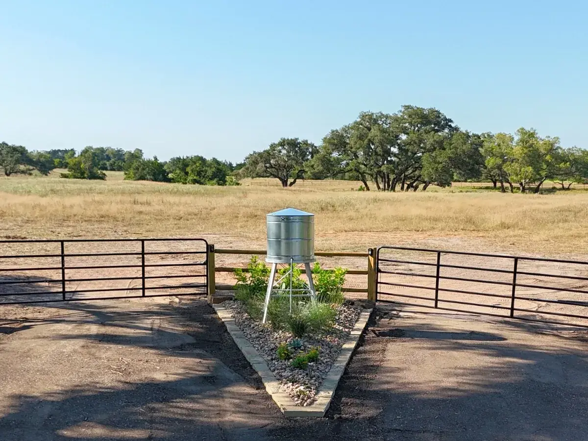 A silver water tank on a stand at the Walker Branch community entrance, framed by black pipe gates, with dry grass and spreading live oaks under a clear sky.