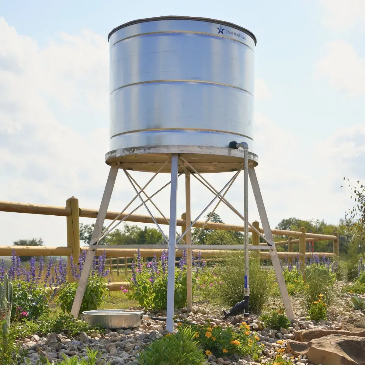 A galvanized water station on a steel stand, with a ground basin and purple salvia blooming around it.